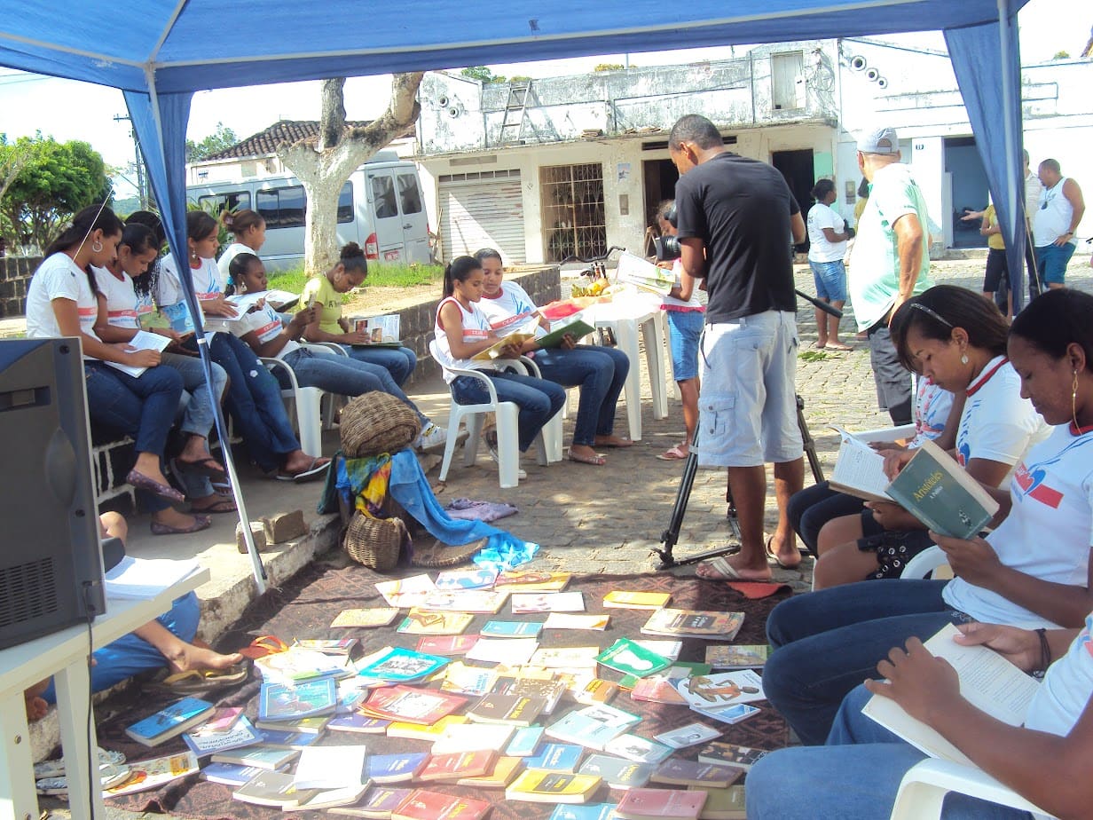 Jovens e adultos lendo sob uma tenda azul ao ar livre, com livros espalhados sobre lonas no chão