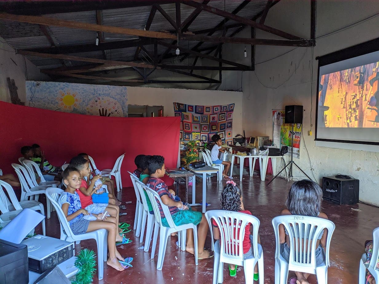 Crianças em cadeiras de plástico assistindo a um filme projetado no salão da ACAPEB