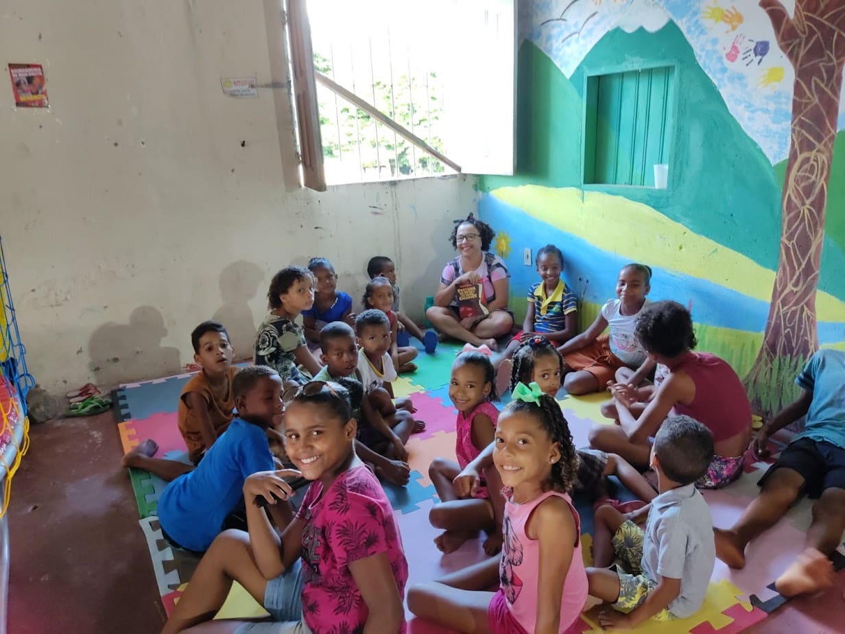Crianças sentadas em tapetes coloridos ouvindo uma contadora de histórias, em sala com parede pintada