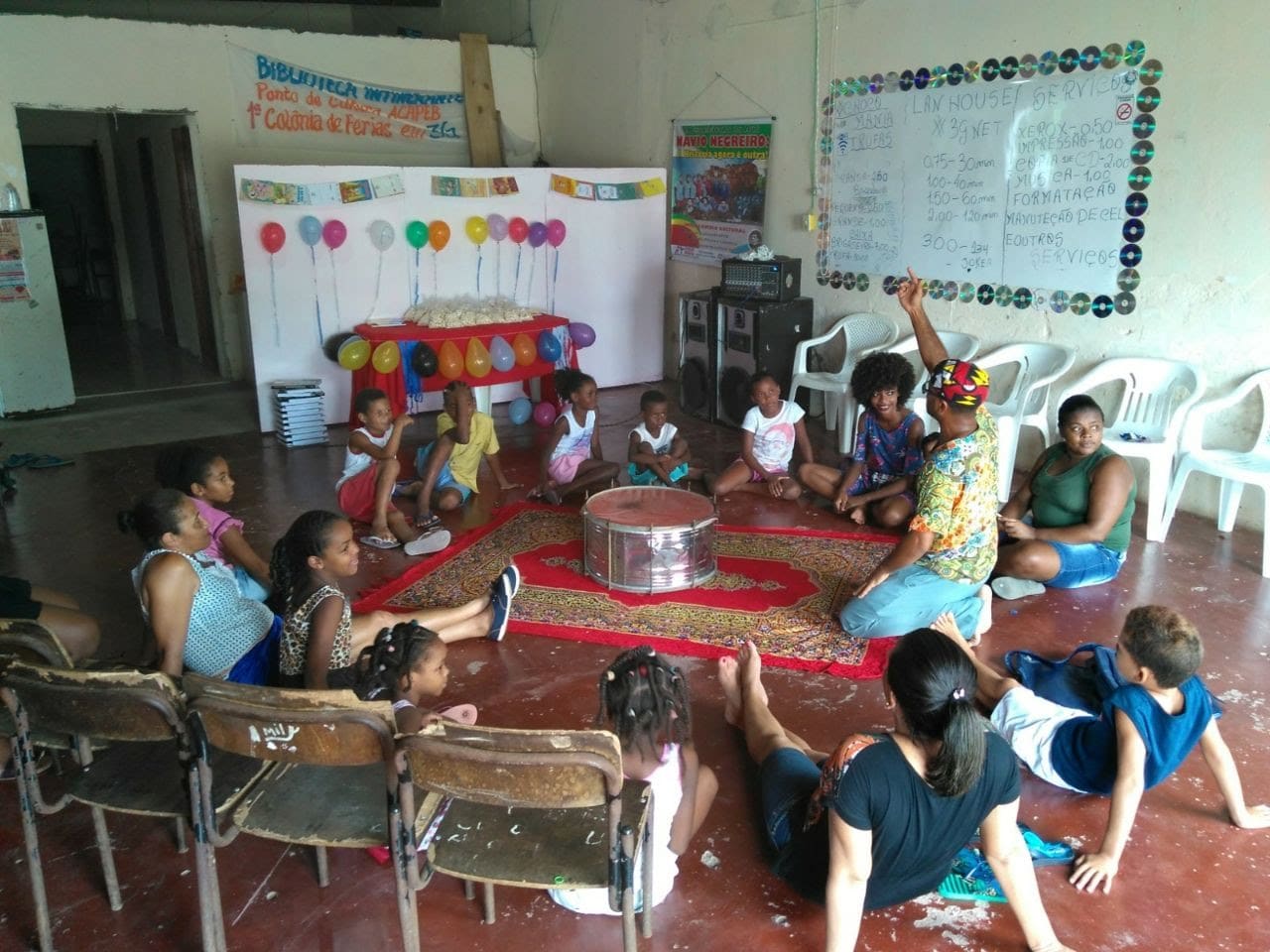 Crianças em roda no chão durante atividade da 1ª Colônia de Férias em 3G, com faixa da Biblioteca Itinerante ao fundo